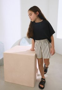 Haut noir à manches courtes, short crème rayé et sandales noires ; debout à côté d'un piédestal géométrique rose clair dans un espace lumineux.