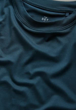 Tumma teelinvihreä kangas, sileä pinta, kerrostettu laskoksin. Pyöreä pääntie, jossa kauluksen lähellä teksti "THE SET" sisältävä merkki.