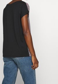 Svart kortärmad topp med mönstrade sidopaneler i röd, vit och marinblå färg. Tyget är mjukt med en avslappnad passform och rundad halsringning.