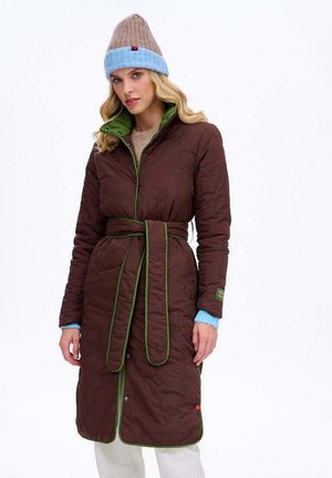 Brązowy pikowany długi płaszcz z zielonym wykończeniem, z paskiem w talii i bocznymi kieszeniami; nałożony na beżowy sweter, w zestawieniu z niebiesko-brązową dzianinową czapką.