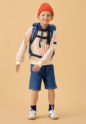 Young boy smiling, wearing red beanie, striped long-sleeve shirt, denim shorts, sneakers, and a blue backpack with chest straps against beige background.