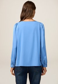 Hellblaue Bluse mit langen Satinärmeln, die mit gerafften Schulterdetails und knopfartigen Manschetten versehen ist, kombiniert mit dunklen Jeans.
