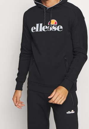 Mężczyzna noszący czarną bluzę z kapturem ellesse i pasujące czarne spodnie dresowe z logo na piersi i udzie na jednolitym tle.