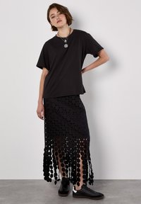 T-shirt in cotone nero, vestibilità comoda. In basso, una gonna maxi nera con ritagli circolari e orlo a frange sovrapposte, indossata con sneakers nere.