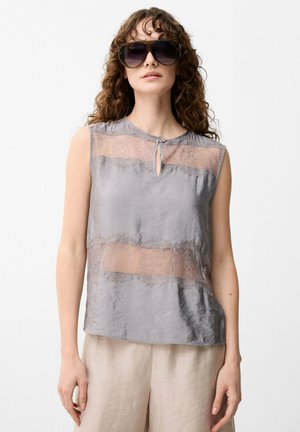 Femme aux cheveux bouclés portant de grandes lunettes de soleil foncées, un top gris sans manches en dentelle et satin, et un pantalon beige sur un fond uni.