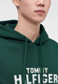 Млад човек, носещ тъмнозелено худи с бял частичен текст "TOMMY HILFIGER", видим отпред.