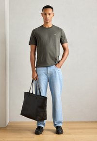 Olive green cotton t-shirt, light blue denim jeans, black leather tote bag with long straps, and black slip-on shoes. Plain background.