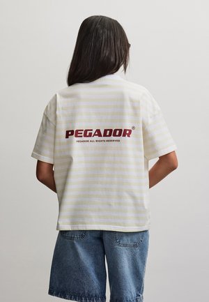 Person med langt mørkt hår iført en lys, stribet T-shirt med teksten "PEGADOR" på ryggen og løse denimshorts, stående mod en ensfarvet baggrund.