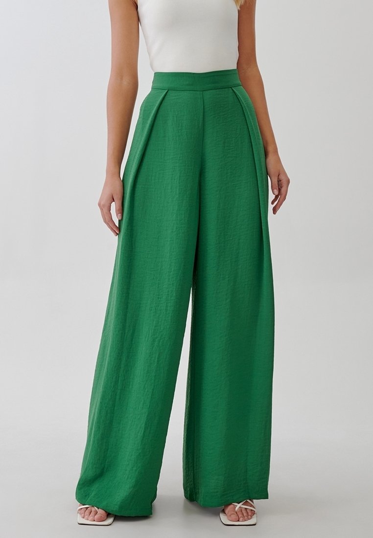 Femme portant un pantalon vert taille haute à jambes larges avec des plis, associé à un haut blanc sans manches et des sandales blanches ouvertes.