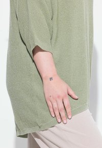 Maglione a maglia verde chiaro, testurizzato, con maniche corte, caratterizzato da una vestibilità ampia e un orlo arrotondato. Tatuaggi visibili sulla mano.