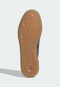 Suela de zapatilla de goma en beige con patrones circulares y texturizados; presenta el logo de la marca. Los bordes laterales se estrechan ligeramente para obtener una forma aerodinámica.