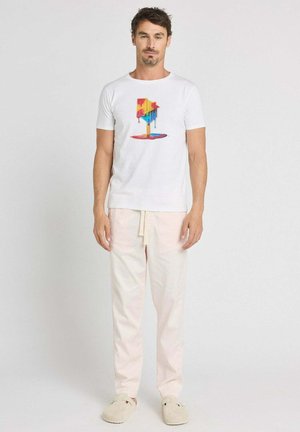 Un homme debout face à l'avant portant un T-shirt blanc avec un motif coloré de boîte aux lettres fondue, un pantalon beige clair et des chaussures beige sans lacets sur un fond uni.