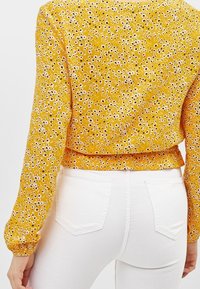 Gul blommig blus med långa ärmar och en insydd midja. I kombination med figursydda vita byxor som har en mjuk, lätt textur.