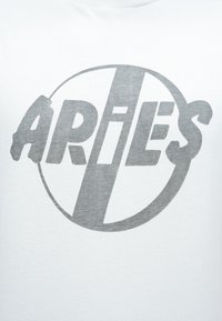 Tela blanca con un texto estilizado en gris y en negrita "ARIES" dentro de un círculo, con una franja diagonal que atraviesa las letras.
