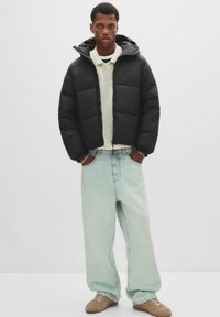 Ung mand iført en sort pufferjakke, en cremefarvet polo-shirt, lyseblå poserede jeans og beige sneakers, der står med hænderne i lommen.