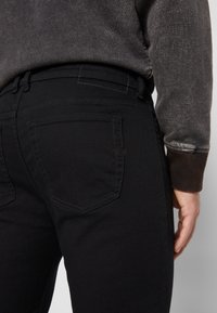 Black jeans made of denim, with a slim fit design, featuring back pockets, subtle stitching, and a smooth texture.