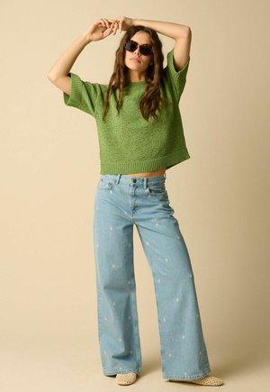 Woman wearing sunglasses, a loose green knit top, light blue wide-leg jeans with small white flower prints, and beige woven shoes, posing with arms raised.