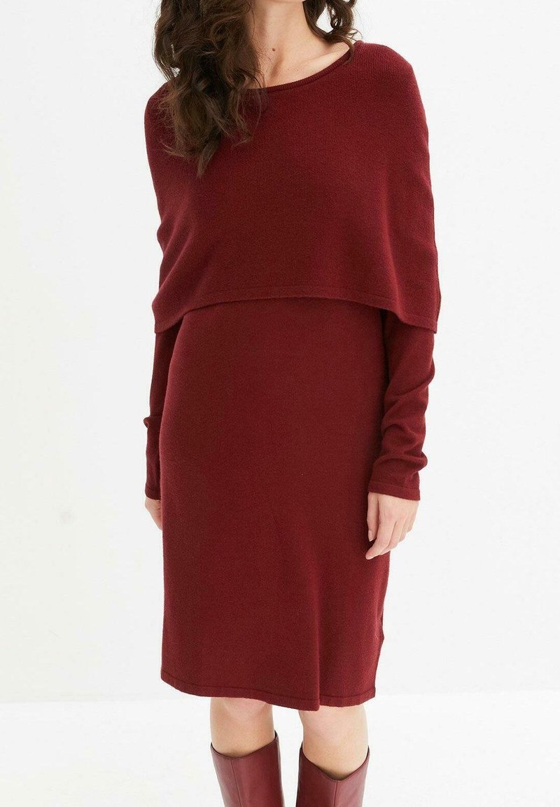 Femme portant une robe bordeaux mi-longue à manches longues avec un châle assorti à couches et des bottes bordeaux mi-mollet, debout devant un fond blanc.