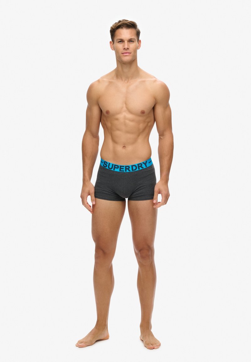 Boxer cortos grises con un logotipo azul de "SUPERDRY" en la cinturilla. Fabricados con tejido suave, diseñados para ofrecer comodidad y un ajuste ceñido.