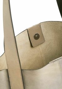 Sac fourre-tout en cuir beige avec un extérieur lisse, doté d'une fermeture à pression et d'une texture intérieure douce semblable au suède. Bandoulière visible en haut.