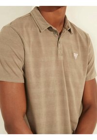 Polo beige en tissu doux, avec un col à boutons, des manches courtes et un petit logo triangulaire sur la poitrine.