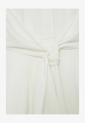Detailný záber na biely textilný odev zviazaný uzlom v páse, zobrazujúci textúru a záhyby materiálu.