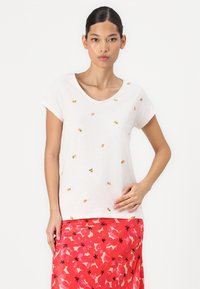 White short-sleeve t-shirt with small orange pineapple embroidery. Smooth texture, scooped neckline, worn with a colorful floral skirt.