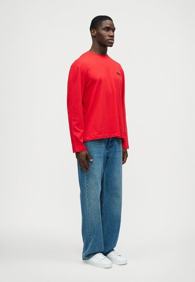 Κόκκινο μακρυμάνικο μπλουζάκι με στρογγυλή λαιμόκοψη, συνδυασμένο με ανοιχτό μπλε χαλαρές τζιν παντελόνες και λευκά αθλητικά παπούτσια. Μινιμαλιστικός σχεδιασμός χωρίς ορατά σχέδια.