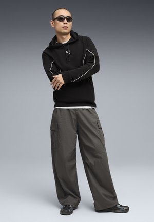 Hombre con sudadera negra con ribete blanco, pantalones cargo gris, zapatillas negras y gafas de sol oscuras, de pie contra un fondo gris.