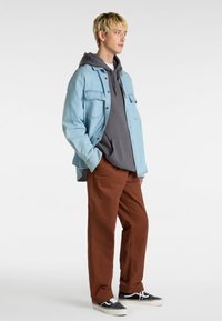 Veste en denim bleu clair sur un sweat à capuche gris, pantalon marron et sneakers noires avec des semelles blanches. Le modèle se tient avec les mains dans les poches.