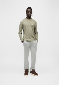 Μπλούζα μακρυμάνικη σε ελιά πράσινο με μικρό λογότυπο, ανοιχτό γκρι υφασμάτινο παντελόνι και καφέ αθλητικά παπούτσια με λευκές σόλες. Το μοντέλο στέκεται με τα χέρια στις τσέπες.