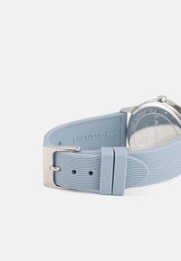 Lacoste kellal on helesinine silikoonist rihm, millel on soonikkoestus ja hõbedane metallist pannal. Ümmargune korpus koos nähtava klambri kujundusega.
