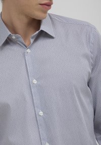 Homme portant une chemise à boutons de couleur claire avec un petit motif géométrique répétitif, col ouvert, regardant légèrement vers la gauche.