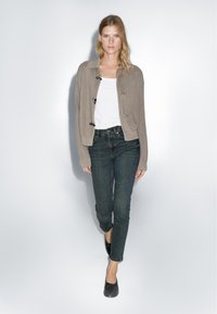 Light brown cropped cardigan with a textured knit, closed with dark buttons, worn over a white top and paired with dark denim jeans.