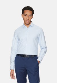 Camisa de cuadros azul claro con cuello estructurado, de manga larga y textura suave. Combinada con pantalones azul marino y un cinturón negro.