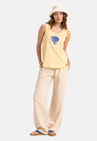 Débardeur jaune avec un graphisme de coquillage bleu, pantalon large crème et tongs bleues. Le mannequin porte également un chapeau seau blanc.