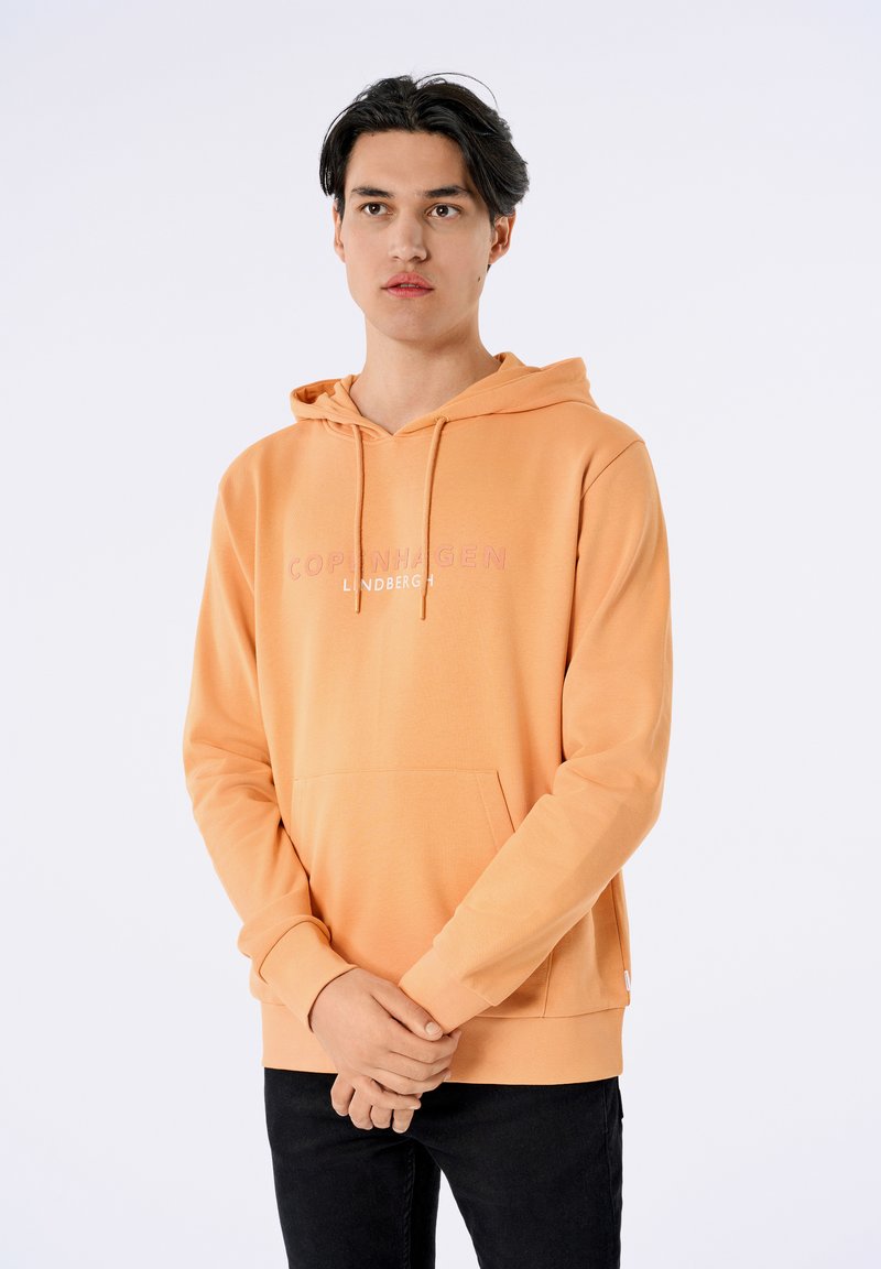 Persikofärgad hoodie med en känguruficka, dragsko i huvan och diskret logotext i ljusrosa centrerad på bröstet. Mjuk textur, avslappnad passform.