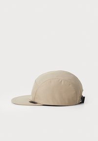 Beige sapka lekerekített felsővel és ívelt szélekkel. Az anyag sima, varrás részletekkel. Hátul egy fekete állítható pánttal rendelkezik.