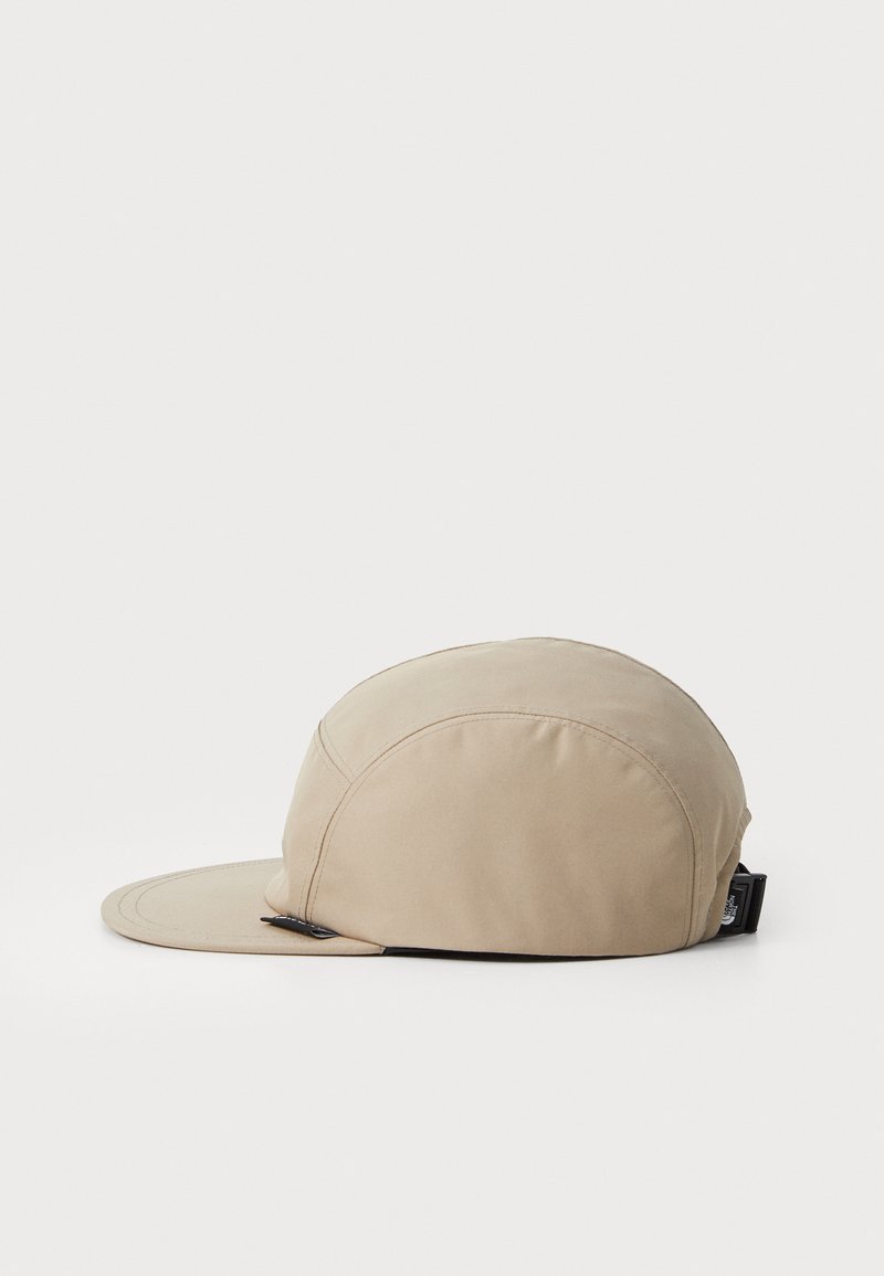 Beige pet met een ronde bovenkant en een gebogen klep. Het materiaal lijkt glad met stikdetails. Voorzien van een zwarte verstelbare band aan de achterkant.