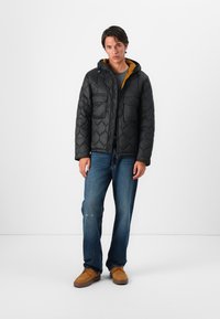 Casaco puff preto acolchoado com capuz, duas bolsos frontais, material leve, usado sobre uma camisa cinza, combinado com jeans azuis e sapatos castanhos.
