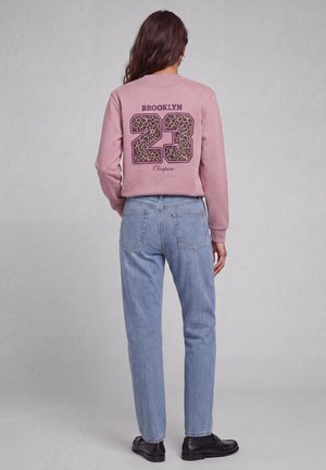 Mujer con cabello largo y castaño que lleva una sudadera rosa con "BROOKLYN 23 Champions" y números con estampado de leopardo, vaqueros azul claro y zapatos negros.