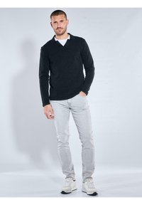 Schwarzer langärmeliger V-Ausschnitt-Pullover, hellgraue Jeans und weiße Sneaker. Das Modell steht vor einem hellen Hintergrund mit neutralem Gesichtsausdruck.