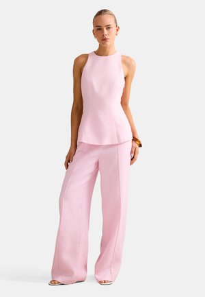 Vrouw draagt een mouwloze lichtroze top met wijde, bijpassende broekspijpen, staat met één hand in de zak en de andere langs haar zijde.