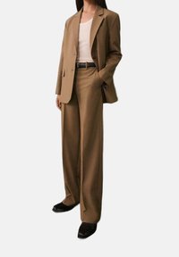 Femme portant un costume marron avec un pantalon à jambes larges, un blazer assorti, un débardeur blanc, une ceinture noire et des chaussures noires à bouts pointus.