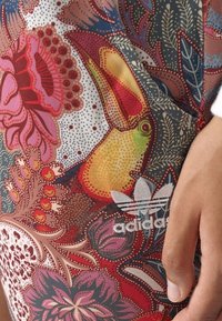 Barevné legíny z látky s potiskem, které mají květinový vzor s tukánem, doplněné bílým logem Adidas na spodní části nohy.