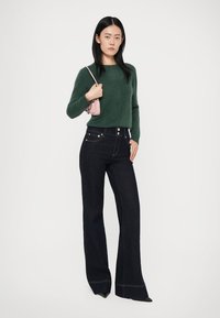 Grüner Wollpullover kombiniert mit dunklen, ausgestellten Jeans mit fünf Taschen und Kontrastnähten. Model hält eine kleine rosa Handtasche.