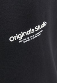 Tessuto nero con finitura testurizzata, caratterizzato da testo ricamato in bianco: "Originals Studio" e ulteriore branding sottostante.