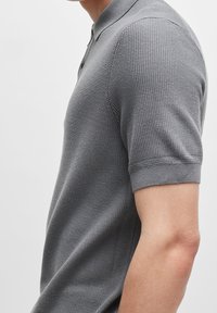 Šedé polo triko s texturovanou tkaninou, krátkým rukávem a límečkem. Má tři knoflíky na límci a přiléhavý střih.