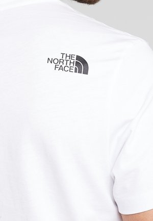 Biała koszula noszona przez osobę z logo "The North Face" wydrukowanym na czarno na górnej części pleców w pobliżu ramienia.