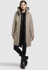 Beige waterdichte jas met een capuchon, voorrits, zijzakken en zwarte accenten. Soepele textuur, knielange ontwerp. Draag over zwarte kleding.